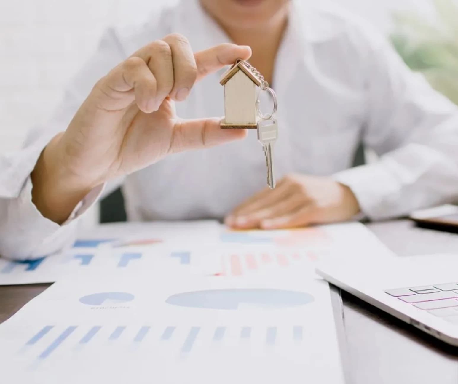 Real estate professional holding house keys above property analytics documents and laptop on a desk